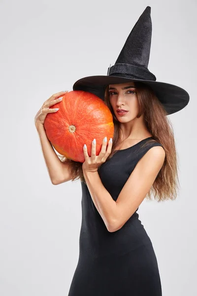 Krásné, kouzelné čarodějnice dívka v klobouku, držící dýně na šedém pozadí. Koncept Halloween. — Stock fotografie