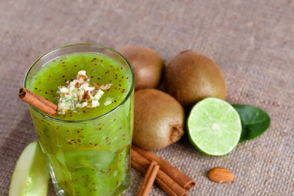 Close-up of a blended drink from ripe kiwi and lime with grated almonds and cinnamon sticks on a light brown fabric background. A few whole juicy kiwi, lime cut in half and a slice of apple on a desk.