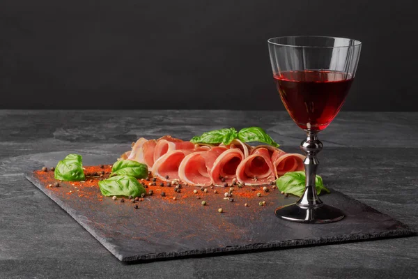 Einen in dünne Scheiben geschnittenen Balyk und ein Glas Rotwein auf dem gesättigten schwarzen Hintergrund. trockener Rotwein, traditionelle italienische Snacks, Basilikum und Pfefferkörner auf dem Tisch. Kopierraum. — Stockfoto