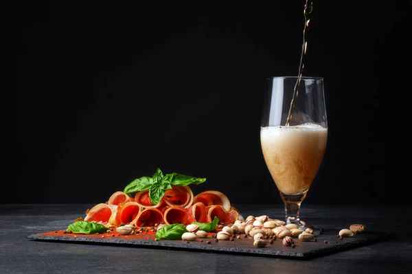 Rohschinken mit Basilikum und einem Becher alkoholischen Bieres mit Schaum auf schwarzem Hintergrund. salzige Nüsse, schmackhafte Schinkenscheiben. und ein Glas Bier. Kopierraum. — Stockfoto