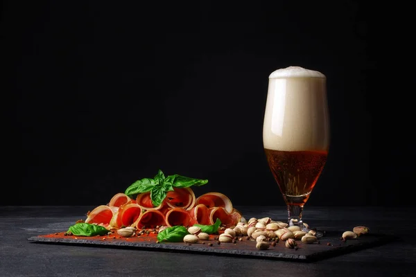 Rohschinken mit Basilikum und einem Becher alkoholischen Bieres mit Schaum auf schwarzem Hintergrund. salzige Nüsse, schmackhafte Schinkenscheiben. und ein Glas Bier. Kopierraum. — Stockfoto