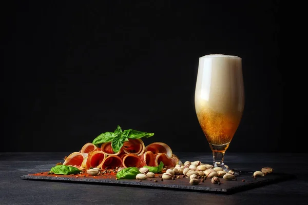 Rohschinken mit Basilikum und einem Becher alkoholischen Bieres mit Schaum auf schwarzem Hintergrund. salzige Nüsse, schmackhafte Schinkenscheiben. und ein Glas Bier. Kopierraum. — Stockfoto