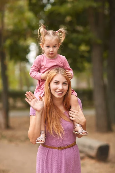 Uma pequena filha senta-se em uma mãe amigável em seus ombros passeando no parque . — Fotografia de Stock