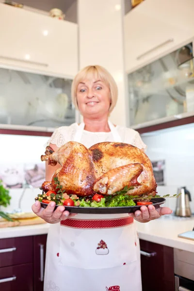 Happy woman holding garnished roasted turkey on a kitchen background. Thanksgiving turkey concept. Copy space.