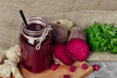 Pancar suyu ahşap arka plan üzerinde siyah saman ile bir mason kavanoz close-up. Bütün ve kesme beetroots, zencefil ve maydanoz kumaş üzerinde. Bir kesme tahtası üzerinde yemek pişirmek için sağlıklı malzemelerle bir sürü.