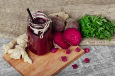 Bir görüntü yukarıdan saman ve birçok organik maddeler ile kırmızı içecek bir büyük mason kavanoz bir kesme tahtası ve ahşap arka plan. Bütün ve kesme beetroots, zencefil ve maydanoz kumaş üzerinde.