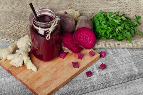 A view from above of a big mason jar of red beverage with straw and many organic ingredients on a cutting board and on a wooden background. Whole and cut beetroots, ginger and parsley on a fabric.