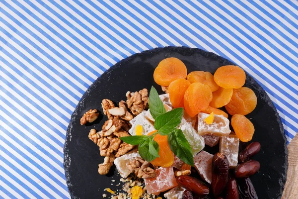 Yakın çekim resmini farklı şekerlemeler malzemelerle çizgili kumaş arka plan üzerinde siyah bir tabak. Turuncu kurutulmuş kayısı, besleyici ceviz, nane yaprağı, Kuru tarihi meyve, Türk lokumu.