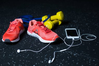 Kadın, beyaz cep telefonu kulaklık, iki sarı canlı dambıl, koyu mavi genişletici, karanlık bir arka plan bulanık egzersiz programı için donatım için koyu kırmızı parlak spor ayakkabı.