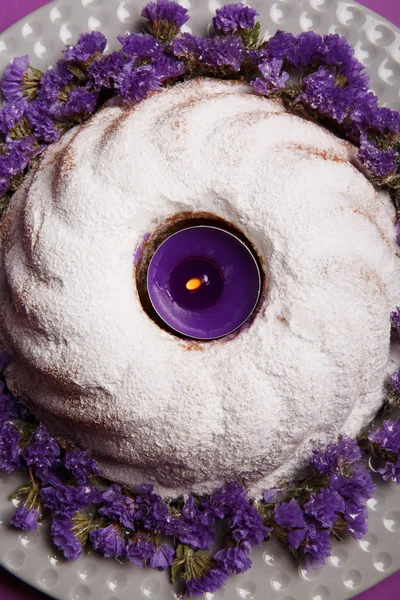 Close-Up bir plaka ile yuvarlak bir kek, pudra şekeri ve yanan bir mor mum şehir merkezinde küçük çiçekler parlak mor arka plan, üstten görünüm üzerinde twigs ile serpilir. Yeni yıl, Noel kavramı.