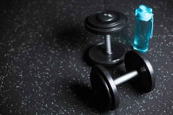 Bir mavi plastik şişe su, iki ağır metal dambıl, spor karşılaşmaları, spor salonları, eğitimler, karanlık bir arka plan bulanık rutinleri için donatım.