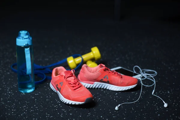 Pink bright sneakers for jogging, a white mobile phone with headphones, two yellow dumbbells and a blue bottle for water, equipment for sports routine on a dark blurred background.