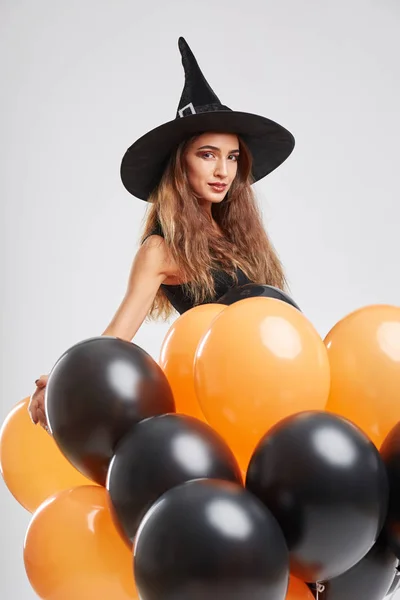 Sexy meisje in een heks kostuum met zwart en oranje ballonnen op een grijze achtergrond. Halloween heks concept. — Stockfoto