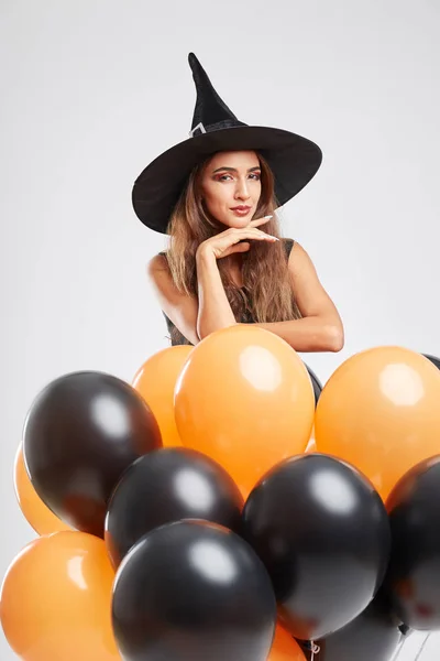 Sexy meisje in een heks kostuum met zwart en oranje ballonnen op een grijze achtergrond. Halloween heks concept. — Stockfoto