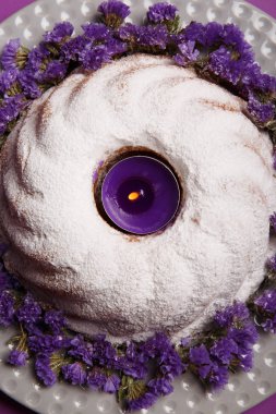 Close-Up bir plaka ile yuvarlak bir kek, pudra şekeri ve yanan bir mor mum şehir merkezinde küçük çiçekler parlak mor arka plan, üstten görünüm üzerinde twigs ile serpilir. Yeni yıl, Noel kavramı.