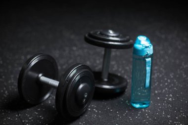 Bir mavi plastik şişe su, iki ağır çelik dambıl, ağırlık çalışması, egzersiz güreşçiler, vücut geliştiriciler ve spor oyuncular üzerinde karanlık bir arka plan bulanık tarafından kullanılan ekipman için.