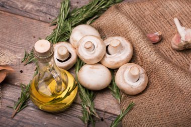 Şeffaf şişe dolu güzel kokulu zeytinyağı, sarımsak ve dallar yeşil biberiye üstten görünüm ahşap arka plan üzerinde. Ormandaki bir çanta ve masanın üzerinde bir kaç taze ve organik mantar.