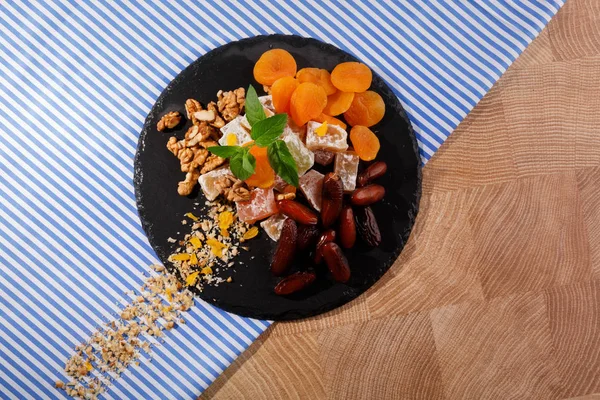 A top view of a black plate full of yummies and fruits on a colorful fabric background. Sugary turkish delight with red date fruits, aromatic mint, dried apricots and crunchy walnuts. Copy space.
