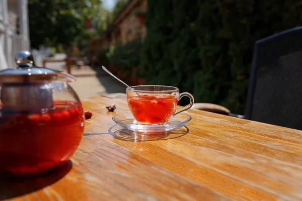 木製テーブルの上の赤いハーブティーの完全美しいカップ ケトル 日当たりの良い通り背景をぼかした写真の茶碗のイチゴ紅茶 外のカフェでイチゴと紅茶 コピー スペース — ストック写真