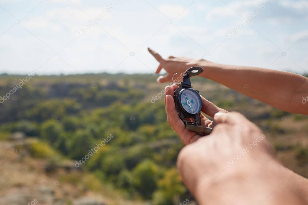 Tourists's hands with compass at mountain road at sunset sky. Close-up view of hand showing little round compass to camera, with incognito man. Point of view shot.