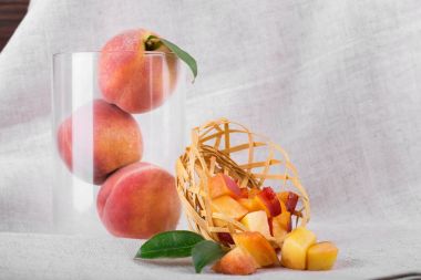 Bir gri kumaş zemin üzerine yeşil yaprakları ile olgun şeftali bileşimi. Bir close-up bir sepet kesme şeftali dolu yanındaki şeffaf bir vazoda üç tüm meyve. Tropikal meyveler tatlılar için.