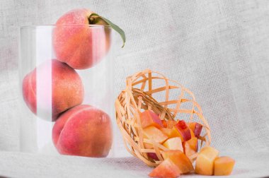 Bir gri kumaş zemin üzerine yeşil yaprakları ile olgun şeftali bileşimi. Bir close-up bir sepet kesme şeftali dolu yanındaki şeffaf bir vazoda üç tüm meyve. Tropikal meyveler tatlılar için.