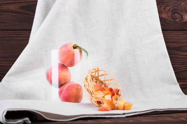 Bir close-up bir sepet kesme şeftali dolu yanındaki şeffaf bir vazoda üç tüm meyve. Koyu ahşap zemin üzerine yeşil yaprakları ile olgun şeftali bileşimi. Tropikal meyveler tatlılar için.