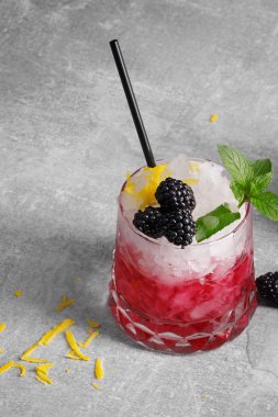Süslü cam soğuk, buzlu içecek dolu parlak nane, limon kabuğu, sulu böğürtlen ve gri taş zemin üzerine siyah bir saman ile. Meyve ve çilek ile kokteyl serinletici. Kopya alanı.