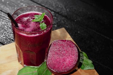 Koyu ahşap arka plan üzerinde siyah bir saman ile tam doğal sebze suyu büyük bir cam. Pancar ve yeşillik kesme masanın yanındaki bir meyve güler yüzlü. Nane baharatlar ile sıkılmış sebze.