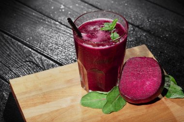 Koyu ahşap arka plan üzerinde siyah bir saman ile tam doğal sebze suyu büyük bir cam. Pancar ve yeşillik kesme masanın yanındaki bir meyve güler yüzlü. Nane baharatlar ile sıkılmış sebze.