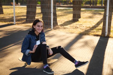 Bir kız parkta bir kaykay üzerinde oturuyor.