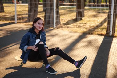 Bir kız parkta bir kaykay üzerinde oturuyor.