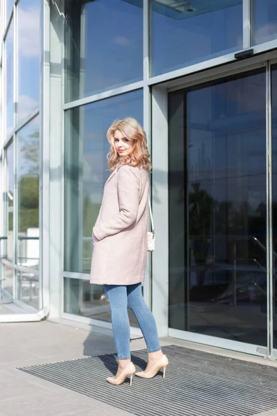 Güzel sarışın genç kız güzel bej bir palto, kot pantolon ve yüksek topuklu.