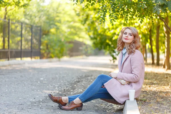 Linda loira está sentada no passeio no parque com café . — Fotografia de Stock