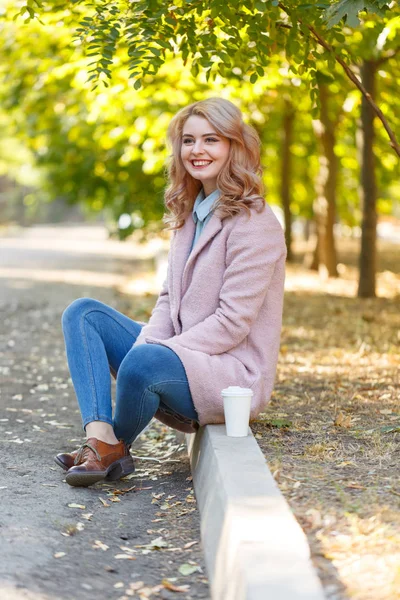 Красива блондинка сидить на бордюрі в парку з кавою . — стокове фото