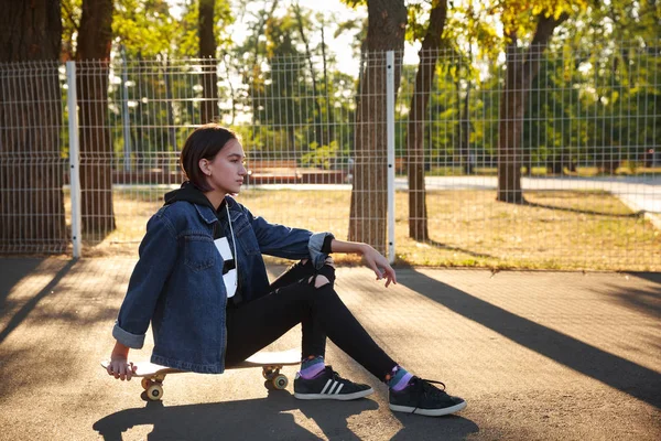 Una chica está sentada en un monopatín en el parque . — Foto de Stock