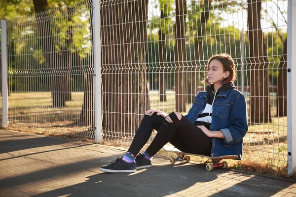 Bir kız parkta bir kaykay üzerinde oturuyor.