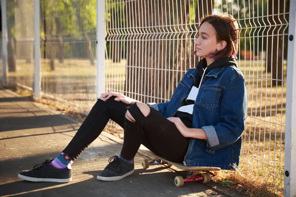 Bir kız parkta bir kaykay üzerinde oturuyor.