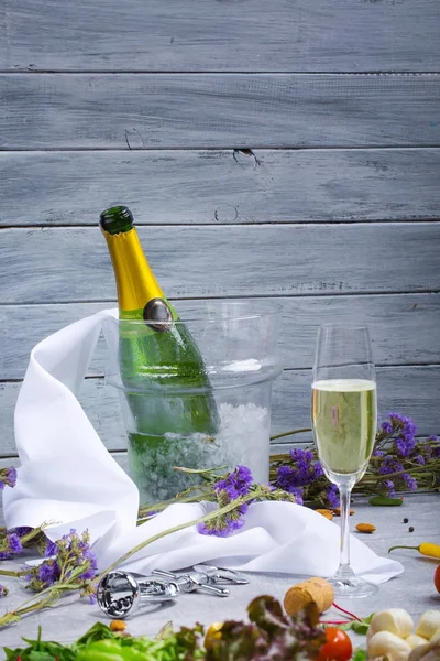 一个玻璃桶与冰和绿色瓶香槟 一杯饮料 金属螺旋状 白色餐巾 小紫罗兰花 木墙上轻木背景 — 图库照片