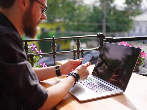 En man sitter på ett träbord håller en telefon. På skrivbordet är en bärbar dator. — Stockfoto