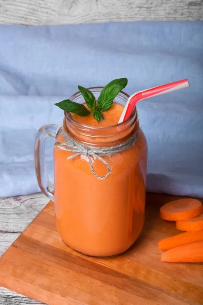 Bir mason kavanoza bir detoks içecek tam bir makro resim. Mavi kumaş arka plan üzerinde serinletici bir havuç yüzlü. Olgun havuç tüpler ve yeşil fesleğen yaprakları kesme masanın yanında bir vitamin içeceği.