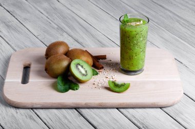 Kivi meyve besleyici vitaminler, tarçın ve ceviz ahşap bir masa ve bir gri tablosudur arka plan dolu. Bir bardağın dolu güler yüzlü sulu kivi, rendelenmiş fındık ve nane bir kesme tahtası üzerinde.