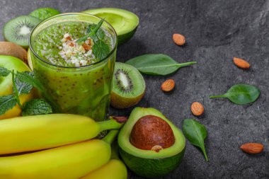Gri taş zemin üzerine farklı egzotik meyvelerin yakın çekim. Muz, besleyici avokado, sulu kivi ve nane bir yığın. Kivi içecek baharat ve fındık ile uzun bir cam.