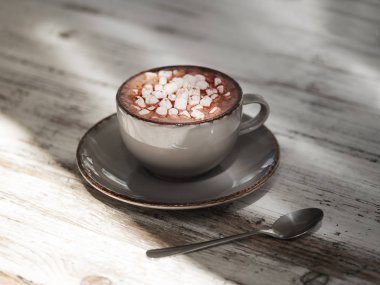 Lezzetli kapuçino gri ahşap masa arka plan üzerinde. Bir gri porselen kupa beyaz marshmallow ile tatlı kakao bardak tam bir makro resim. Metal bir kaşık ile bir fincan. Kahvaltı konsepti.