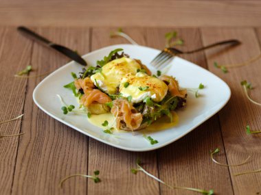 İştah açıcı bir kompozisyon benedict yumurta ile yeşil ve mor salata yaprakları, mayo ve Somon ızgara tost çatal ve bıçak ile bir tabakta et. Bir ahşap masa arka plan üzerine haşlanmış yumurta.