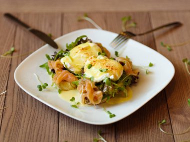 İştah açıcı bir kompozisyon benedict yumurta ile yeşil ve mor salata yaprakları, mayo ve Somon ızgara tost çatal ve bıçak ile bir tabakta et. Bir ahşap masa arka plan üzerine haşlanmış yumurta.