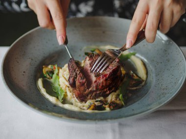 İki el bir tabak içinde sığır eti parçası kesiyorsun. Bir mavi yuvarlak tabak dolusu beyaz masa arka plan üzerinde lezzetli yemeğin bir yakın çekim resim. Lezzetli pahalı yiyecek. Restoran, yiyecek konsepti.