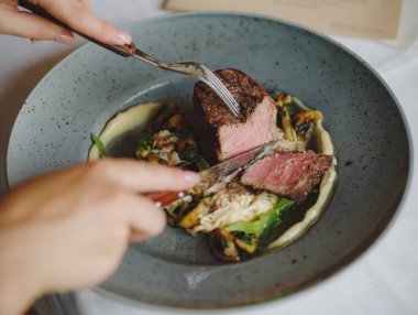 İki el bir tabak içinde sığır eti parçası kesiyorsun. Bir mavi yuvarlak tabak dolusu beyaz masa arka plan üzerinde lezzetli yemeğin bir yakın çekim resim. Lezzetli pahalı yiyecek. Restoran, yiyecek konsepti.