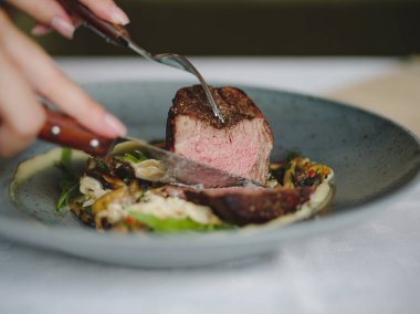 İki el bir tabak içinde sığır eti parçası kesiyorsun. Bir mavi yuvarlak tabak dolusu beyaz masa arka plan üzerinde lezzetli yemeğin bir yakın çekim resim. Lezzetli pahalı yiyecek. Restoran, yiyecek konsepti.