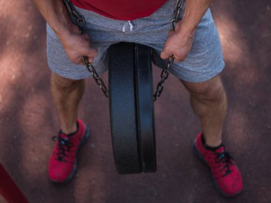 Seksi vücut ile güçlü bir adam kendi ayakları üzerinde egzersizleri yapıyor. Yakışıklı bir genç adam parkta ayaklarıyla ağırlık kaldırıyor.